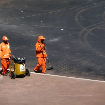 Dois trabalhadores da limpeza urbana usam uniformes laranja enquanto trabalham em Brasília.