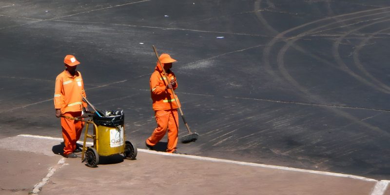 Dois trabalhadores da limpeza urbana usam uniformes laranja enquanto trabalham em Brasília.