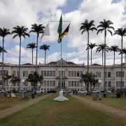 Edifício público em Juiz de Fora, cercado por palmeiras e com bandeiras brasileiras, sob céu nublado.