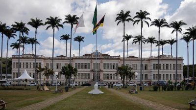 Edifício público em Juiz de Fora, cercado por palmeiras e com bandeiras brasileiras, sob céu nublado.