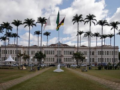 Edifício público em Juiz de Fora, cercado por palmeiras e com bandeiras brasileiras, sob céu nublado.