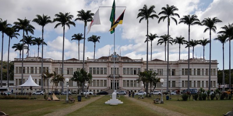 Edifício público em Juiz de Fora, cercado por palmeiras e com bandeiras brasileiras, sob céu nublado.
