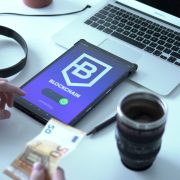 Tablet exibindo uma transação de moeda digital em blockchain ao lado de um laptop e cédulas de euro.
