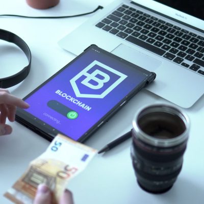 Tablet exibindo uma transação de moeda digital em blockchain ao lado de um laptop e cédulas de euro.