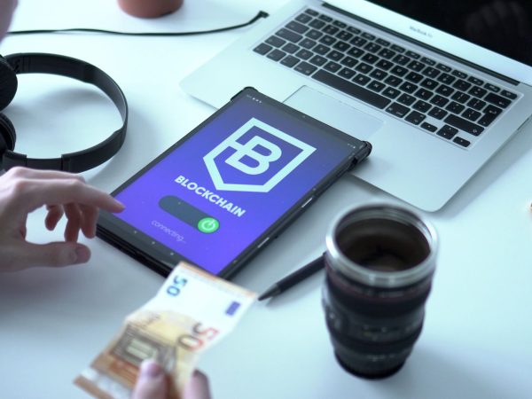Tablet exibindo uma transação de moeda digital em blockchain ao lado de um laptop e cédulas de euro.